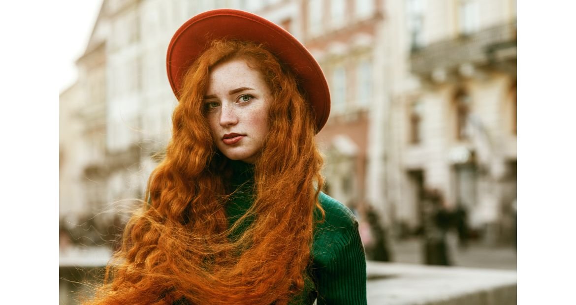Mulher com blusa verde, chapéu vermelho e cabelo ruivo alaranjado.